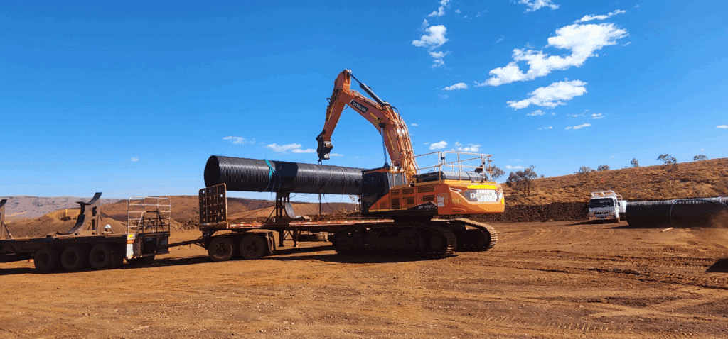 Develon excavator with Gajawari branding unloading drainage pipe from flatbed truck on Western Australia civil construction site for infrastructure projects