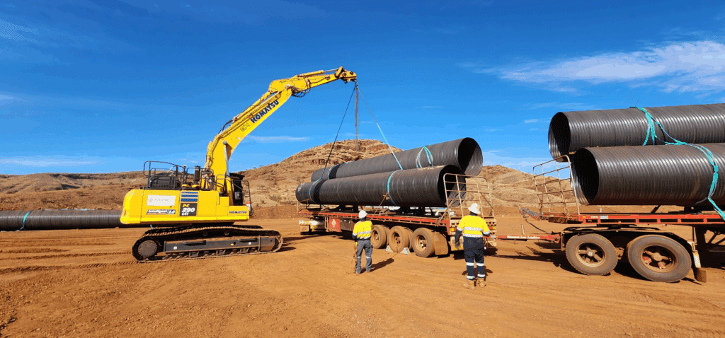 Excavator lifting drainage pipe from truck with construction workers assisting on Western Australia civil site for industrial projects